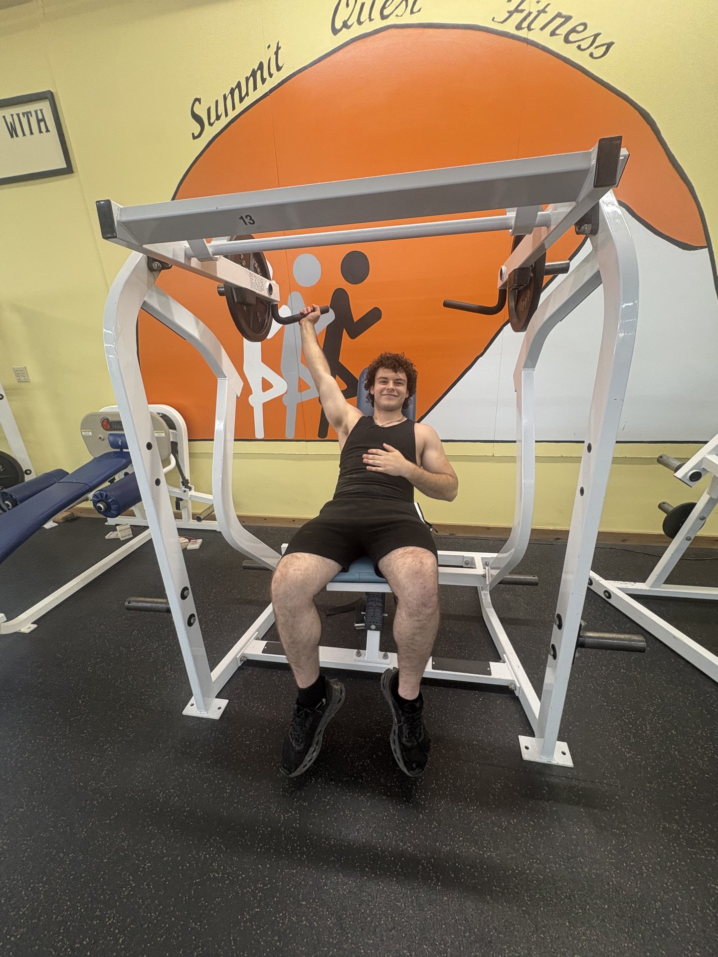 Member training on a plate-loaded press beneath the Summit Quest sunrise mural